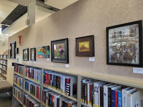 Six framed paintings hung on a wall above a library bookshelf.