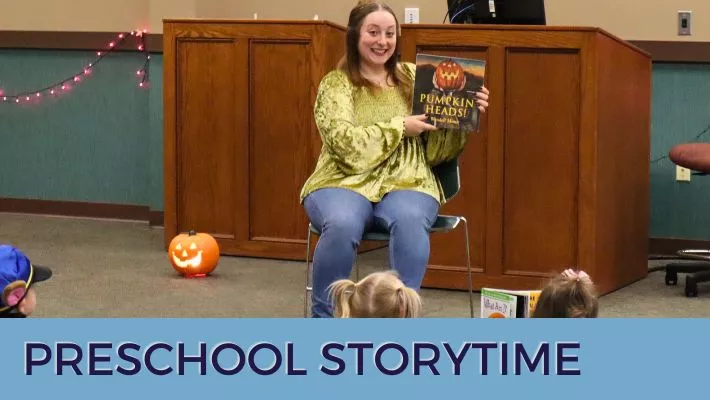 Miss Maddie smiles and holds up a book to a group of children at storytime.